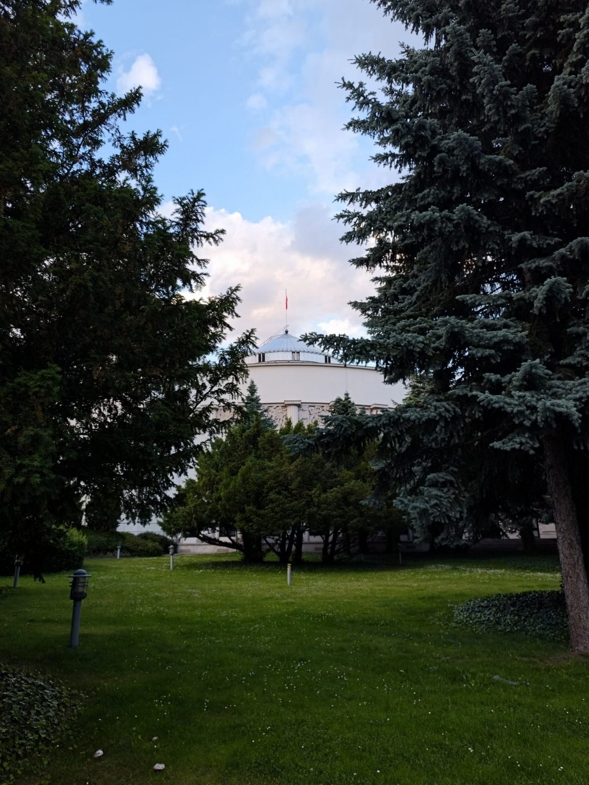 Budynek Sejmu RP z kopułą i flagą, częściowo widoczny między drzewami